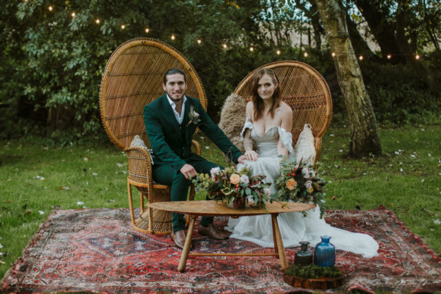 Married couple sitting in boho wooden chairs with flowers on table outdoors